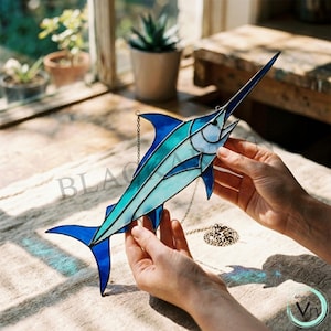 May include: A stained glass marlin in shades of blue and teal, with a long, pointed bill and a chain for hanging. The fish is held by hands, showcasing the intricate details of the glasswork. The design features geometric shapes and lines.