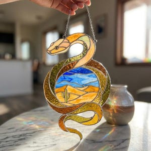 Könnte beinhalten: Buntglas-Schlange mit einer Wüstenlandschaft. Die Schlange ist in Gelb-, Orange- und Brauntönen gehalten, mit einer Wüstenlandschaft in der Mitte. Die Wüstenlandschaft zeigt einen blauen Himmel, Sanddünen und einen Kaktus. Sie hängt an einer silbernen Kette.