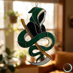 May include: A stained glass suncatcher depicting a hooded figure in a dark green and black robe, holding a long yellow and silver scythe. The figure is wrapped in a green snake-like form, with clear glass accents, all held together by dark metal solder lines.