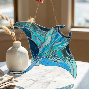 May include: A stained glass manta ray suncatcher in shades of blue and teal, suspended by a gold chain. The artwork features intricate details and a flowing design, creating a beautiful ocean-themed decoration. The suncatcher is held up by a hand.