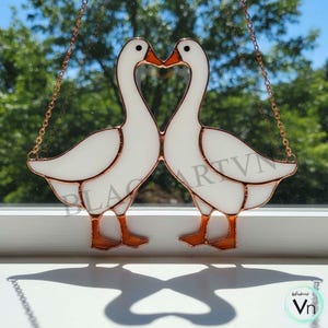 Könnte beinhalten: Glasmalerei mit zwei weißen Gänsen, deren Schnäbel ein Herz bilden. Die Gänse haben orangefarbene Schnäbel und Füße mit kupferfarbenen Umrissen. Das Werk wird von einer Kupferkette gehalten. Der Schatten ist herzförmig.
