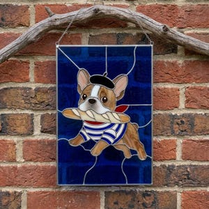 Könnte beinhalten: Ein rechteckiges Buntglasfenster mit einem hellbraun-weißen Französischen Bulldoggen-Motiv, der ein schwarzes Barett und ein blau-weiß gestreiftes Shirt trägt. Der Hund hält ein Baguette im Maul vor einem tiefblauen Hintergrund mit silbernen Metalllötstellen.