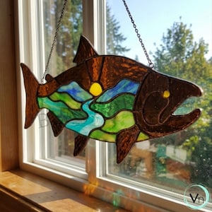 Può includere: Acchiappasole a forma di pesce in vetro colorato con corpo e coda marroni, con un design paesaggistico con un sole giallo, un fiume blu e colline verdi. Il pesce è appeso davanti a una finestra.