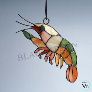 May include: A stained glass shrimp suncatcher with a variety of colors, including orange, green, yellow, and red. The shrimp is hanging from a brown string. The suncatcher has a detailed design and is a decorative item.