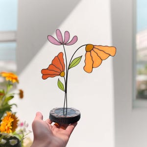 Puede incluir: Una escultura de flores de vidriera con tres flores coloridas. Las flores son en tonos naranja y rosa, con hojas verdes y tallos negros. La escultura está montada sobre una base redonda de color oscuro, sostenida en una mano.
