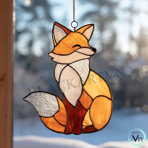 Peut inclure: Suncatcher en vitrail représentant un renard souriant, aux couleurs orange, blanc et marron. Le renard est suspendu à une chaîne. Le renard est composé de plusieurs morceaux de verre.