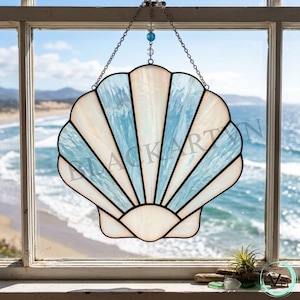 May include: A stained glass suncatcher shaped like a scallop shell, featuring alternating panels of iridescent white and light blue glass. The piece is framed in dark metal solder lines and hangs from a silver chain with a small blue glass bead.