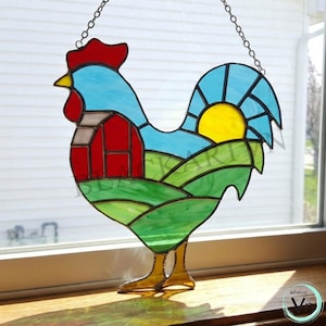 May include: Stained glass rooster suncatcher with a red comb, blue body, and green fields. The design includes a red barn, a yellow sun, and a blue sky. The rooster is hanging from a chain.