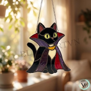 May include: A stained glass suncatcher shaped like a black cat wearing a purple and red vampire cape with a yellow collar. The cat has bright yellow eyes and a white chest patch. It hangs from a silver metal chain against a blurred window background.