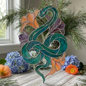 May include: A stained glass artwork depicting a green and teal snake entwined with orange and purple flowers. The piece hangs from a silver chain. The background includes greenery and flowers, creating a vibrant, nature-inspired design.