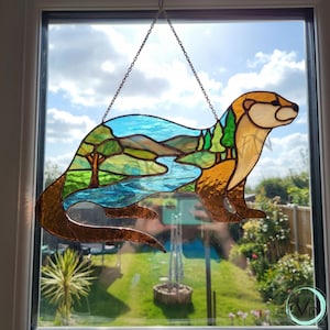 Pode incluir: Decoração de janela de vidro manchado em forma de lontra. A lontra é castanha e bege, com uma paisagem de rio azul e árvores e colinas verdes dentro do seu corpo. Pendura-se de uma corrente em frente a uma janela com um céu ensolarado.