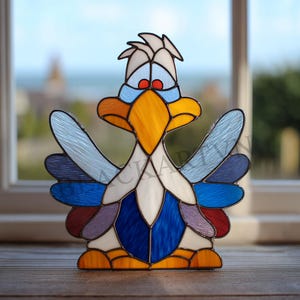 Op de afbeelding: Een gebrandschilderde vogelsculptuur met een cartoonachtig ontwerp. De vogel heeft een gele snavel, rode ogen en vleugels in tinten blauw, paars en wit. De basis is oranje en het stuk staat tegen een vage achtergrond van een raam.