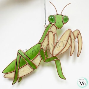 May include: A stained glass praying mantis ornament featuring vibrant green and cream-colored glass panels held together by copper foil solder lines. The insect has two prominent green glass eyes and a thin silver hanging wire attached at the top.