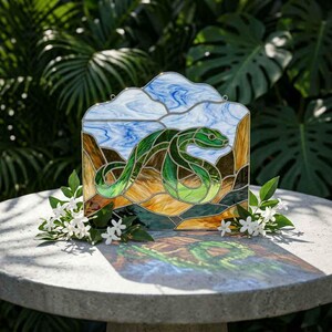 May include: A stained glass panel featuring a green snake coiled against a background of blue and white swirled glass representing the sky and brown textured glass representing the ground, framed by thin metal lines with two small hanging loops.