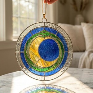 Può includere: Un acchiappasole rotondo in vetro colorato con una catena dorata. Il design presenta un cerchio blu, una mezzaluna gialla e un serpente verde. Il vetro è incorniciato da un bordo trasparente strutturato. L'acchiappasole è tenuto da una mano.