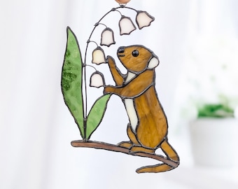 May include: Stained glass art piece featuring a brown and white weasel reaching for white lily of the valley flowers. The artwork includes a green leaf and is outlined with dark lines. The piece is held up by a metal ring.