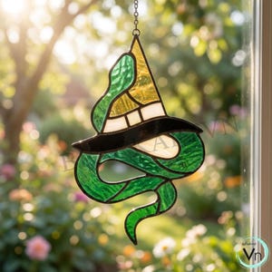 Könnte beinhalten: Ein Buntglas-Suncatcher, der eine grüne Schlange zeigt, die sich um einen gelb-weißen Hexenhut windet. Das Stück ist mit dunklen Metalllötlinien gefertigt, hängt an einer silbernen Kette und zeigt durchscheinende Glasstrukturen und Farben.