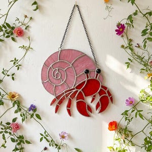 May include: A stained glass hermit crab wall hanging. The crab features a pink spiral shell and a red body with black eyes. The artwork is suspended by a chain and surrounded by climbing vines with flowers.