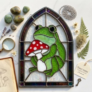 Peut inclure: Un vitrail suspendu représentant une grenouille verte tenant un champignon rouge et blanc. L'œuvre est encadrée dans une arche en métal sombre avec un fond en verre clair texturé. Une petite étiquette attachée indique Forest Friends Stained Glass.