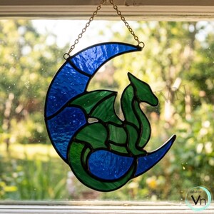 Puede incluir: Atrapasoles de vidriera con un dragón verde dentro de una luna creciente azul. El dragón tiene alas detalladas y una larga cola. El atrapasoles está suspendido por una cadena dorada y se exhibe frente a una ventana.