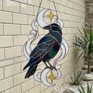 Puede incluir: Decoración colgante de vidrieras con un cuervo. El cuervo está posado en una luna creciente blanca con una estrella amarilla. El cuervo tiene plumas iridiscentes azules y verdes. La decoración está suspendida por una cadena de un gancho de metal negro.