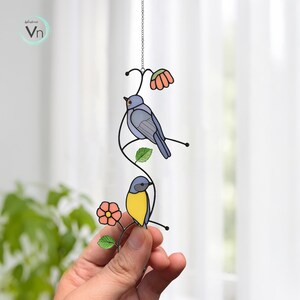May include: A stained glass bird suncatcher featuring two birds perched on a black metal vine with leaves and flowers. The birds are blue and yellow, with orange and green accents. The suncatcher hangs from a silver chain.