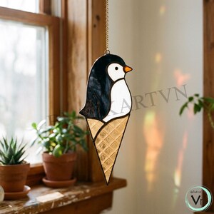 Könnte beinhalten: Glasfenster-Suncatcher eines Pinguins in Form einer Eistüte. Der Pinguin hat einen schwarz-weißen Körper und einen orangefarbenen Schnabel. Die Tüte ist goldfarben und strukturiert. Der Suncatcher hängt an einer Kette.