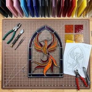 Op de afbeelding: Een glas-in-lood paneel van een feniks in een boogvormig frame met oranje, rode en gele glasstukken. Gereedschap omvat tangen met groene handvatten, een glassnijder, een pincet en tangen met rode handvatten. Een papieren schets en vier vierkante glastegels zijn aanwezig.