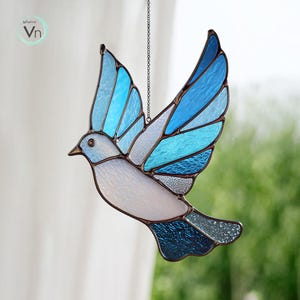 May include: Stained glass bird suncatcher in flight, featuring shades of blue and white. The bird has outstretched wings and is suspended by a chain. The design includes a variety of textures and patterns within the glass, creating a decorative piece.
