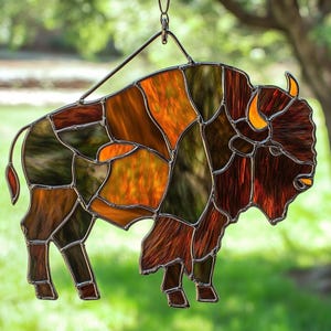 May include: Stained glass bison suncatcher with a metal hanging loop. The bison is composed of various shades of brown, orange, and green glass pieces. The horns are a bright yellow. The suncatcher is hanging in front of a blurred green background.