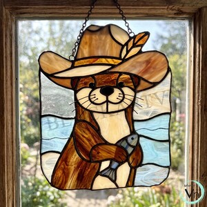 Pode incluir: Um apanhador de sol em vitral representando uma lontra usando um chapéu de cowboy bege com uma pena, segurando um pequeno peixe azul e branco. A lontra tem pelo castanho, peito creme e um fundo de água azul-clara.