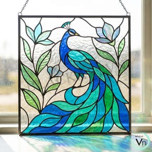 Op de afbeelding: Gebrandschilderd glaspaneel met een pauw in blauwe en groene tinten. Het ontwerp bevat groene en blauwe bladeren, omlijst door een zwart metalen frame. Geschikt voor raam- of wanddecoratie.