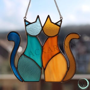 Può includere: Appeso per finestra con gatti in vetro colorato con due gatti stilizzati in verde acqua, arancione e giallo. I gatti sono uno accanto all'altro, con le code ricurve. Il pezzo è incorniciato in metallo scuro e appeso a una catena.