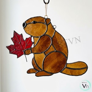 May include: A stained glass suncatcher shaped like a beaver with amber-colored glass pieces held together by dark metal solder lines. The beaver is holding a bright red maple leaf made of textured glass. A small metal loop is attached at the top.