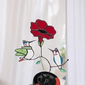 May include: Stained glass hummingbird and flower sculpture. The sculpture features two hummingbirds perched on a branch with a large red flower. The hummingbirds have blue, green, and white details. The flower is red with a black center.