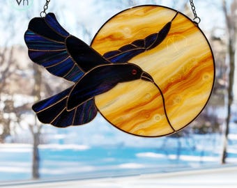 Puede incluir: Vidriera de un cuervo negro en vuelo frente a un sol amarillo y naranja. El cuervo tiene detalles azules y morados y un ojo rojo. El sol tiene un diseño texturizado. La pieza está suspendida por una cadena.