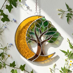 Puede incluir: Atrapasoles de vidriera con un árbol dentro de una luna creciente. La luna es de color amarillo cálido, y el árbol tiene hojas verdes y un tronco marrón. El atrapasoles cuelga de una cadena y está rodeado de vegetación.
