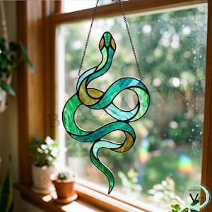May include: A stained glass snake suncatcher in green, blue, and gold, suspended by a chain. The snake design is displayed in front of a window, highlighting the vibrant colours and intricate details. A decorative piece for home decor.