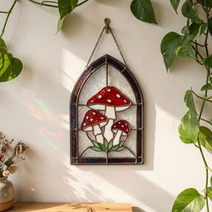 May include: An arched stained glass window hanging featuring three red mushrooms with white spots and white stems. The mushrooms are set against an iridescent, textured glass background, framed in dark metal, and suspended by a metal chain.