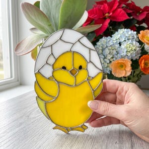 Peut inclure: Décoration de Pâques en vitrail représentant un poussin jaune sortant d'un œuf blanc et jaune. Le poussin a des yeux noirs et des ailes déployées. L'œuf est souligné d'argent. La décoration est tenue devant une fenêtre avec des fleurs en arrière-plan.