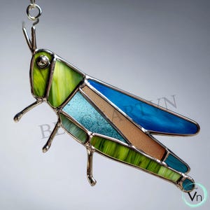 May include: A stained glass grasshopper suncatcher with a silver metal frame. The insect features shades of green, blue, and brown glass. The suncatcher is suspended by a silver chain and is a decorative item.