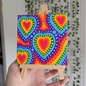May include: A small, colorful canvas painting on a wooden easel. The artwork features a repeating heart pattern in vibrant rainbow colors, including red, orange, yellow, green, blue, and purple. The painting is held by a hand.