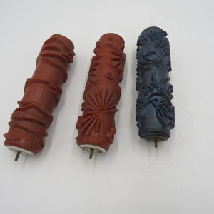 May include: Three paint rollers with different patterns. The roller on the left has a geometric pattern, the middle roller has a fan-shaped pattern, and the roller on the right has a floral pattern. The rollers are all red and blue.