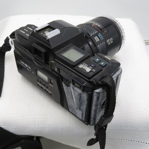 May include: A black Minolta Maxxum 7000 SLR camera with a black Minolta AF lens attached. The camera has a digital display and a black strap.