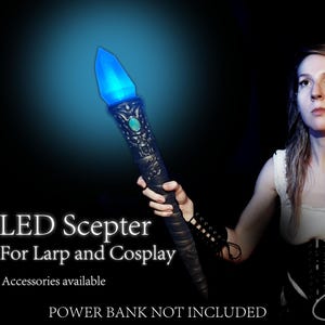 Puede incluir: Un cetro de color oscuro con un mango ornamentado, un acento verde central y una punta de cristal LED azul brillante. El texto en la imagen dice: LED Scepter For Larp and Cosplay, Accessories available, POWER BANK NOT INCLUDED.