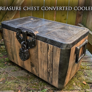 Puede incluir: Nevera rústica diseñada para parecer un cofre del tesoro. Cuenta con un exterior de madera envejecida, una tapa negra texturizada y un cierre metálico decorativo con candado. El texto "TREASURE CHEST CONVERTED COOLER" está en la parte superior.