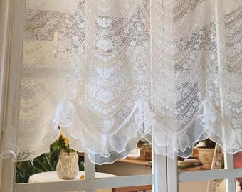 Op de afbeelding: Witte kanten raamgordijn met een geschulpte onderrand. Het gordijn heeft een bloemenpatroon en hangt voor een raam. Het raamkozijn is wit en de achtergrond toont een kamer met een tafel en stoel.