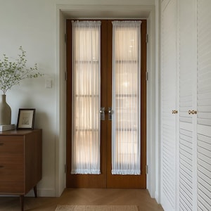 May include: Two sheer white vertical curtains are mounted on a set of double wooden doors with glass panels. The curtains feature a gathered top and bottom hem, covering the glass sections of the doors, which have silver metal door handles.