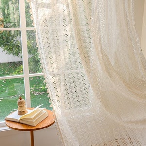 Könnte beinhalten: Weißer Häkelvorhang mit geometrischem Muster und Fransenbesatz. Der Vorhang hängt an einem Fenster und weht im Wind.