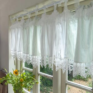 May include: White lace-trimmed window curtains. The curtains are made of a sheer, striped fabric and are hung on a white rod. The bottom edge of the curtains features a delicate lace trim. A vase of yellow flowers sits on the windowsill.